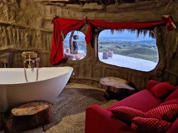 Freestanding tub in a curved stone bathroom with red drapery and a savanna view at Africa Amini Original Maasai Lodge