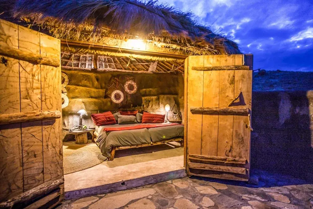 Rustic stone-walled guest bedroom with a carved wooden door at Africa Amini Original Maasai Lodge lit at dusk