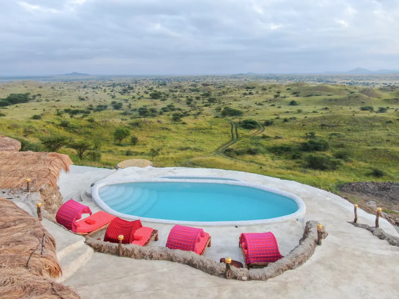 Oval infinity pool with red Maasai-cloth loungers overlooking rolling savanna hills at Africa Amini Original Maasai Lodge
