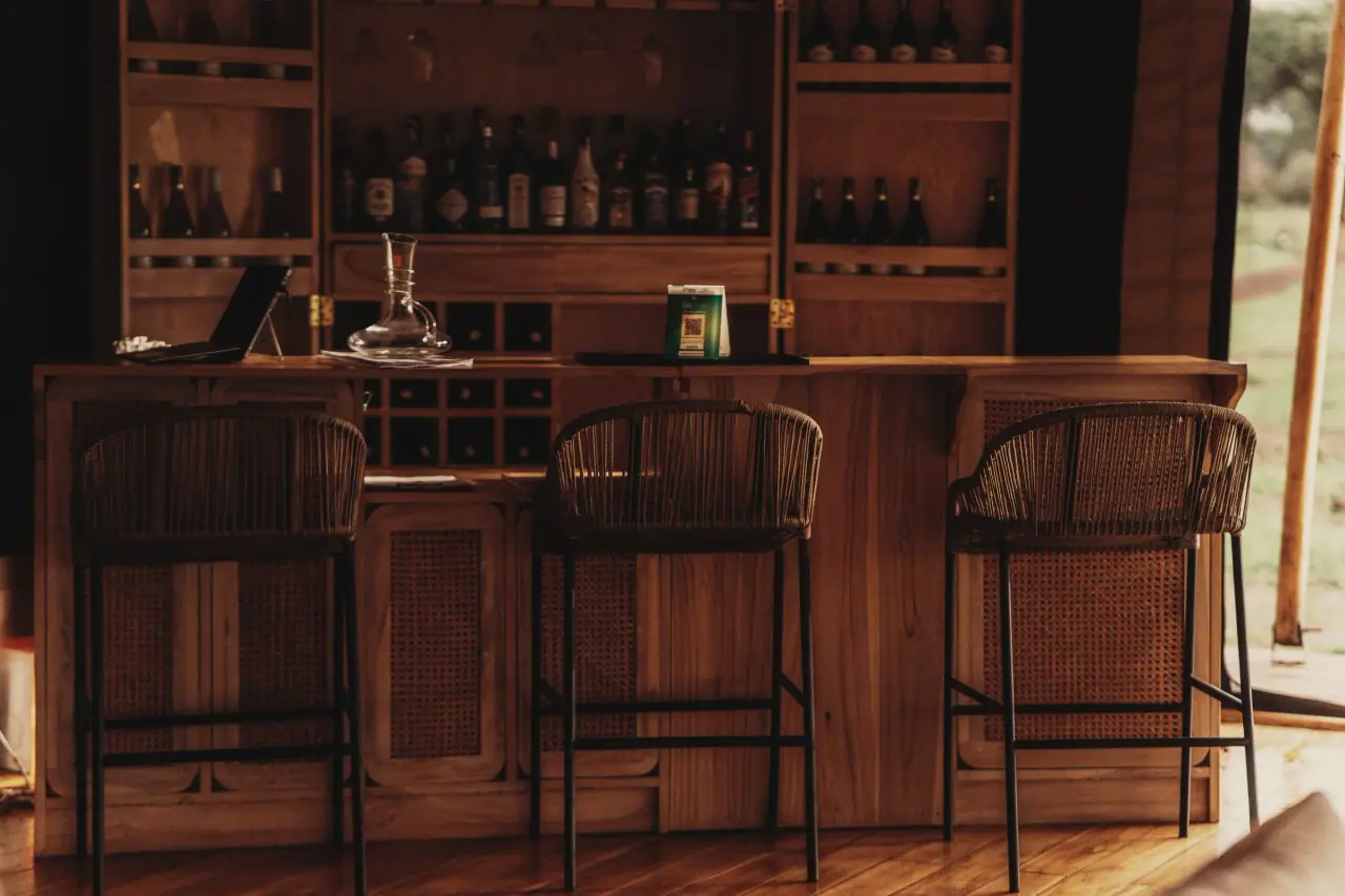 Wooden camp bar stocked with spirits and rattan stools at Conserve Safari Tarangire