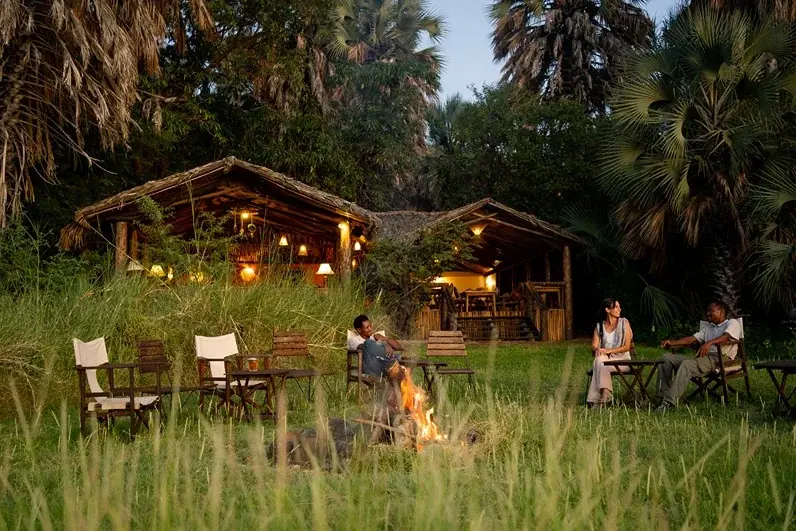 Thatched lounge and dining buildings lit at dusk with guests gathered around a fire pit at Kisima Ngeda Tented Camp