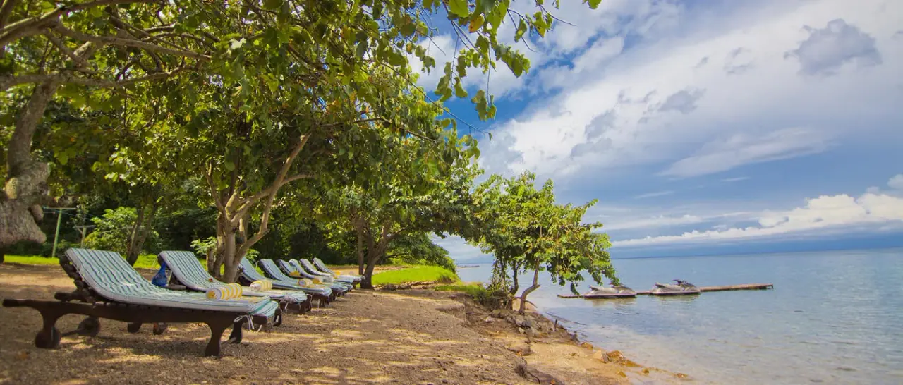 Sun loungers shaded by trees on the Lake Tanganyika shoreline at Mbali Mbali Gombe Tented Camp
