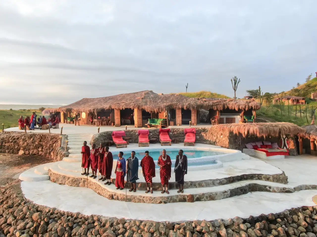 Maasai warriors in traditional shuka gathered around the pool at Original Maasai Lodge with the thatched lodge behind