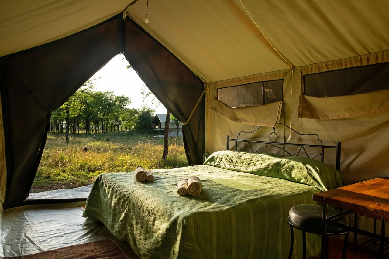 A double bed with green linen and an open tent flap onto the bush at Tarangire Katikati Tented Camp