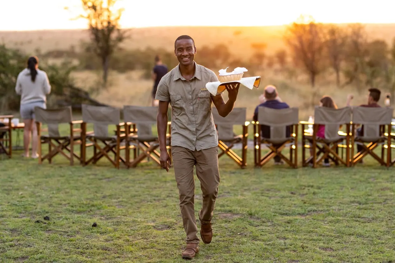 A guide carrying a tray of drinks across the lawn as guests sit in director chairs at sunset at Tarangire Katikati Tented Camp