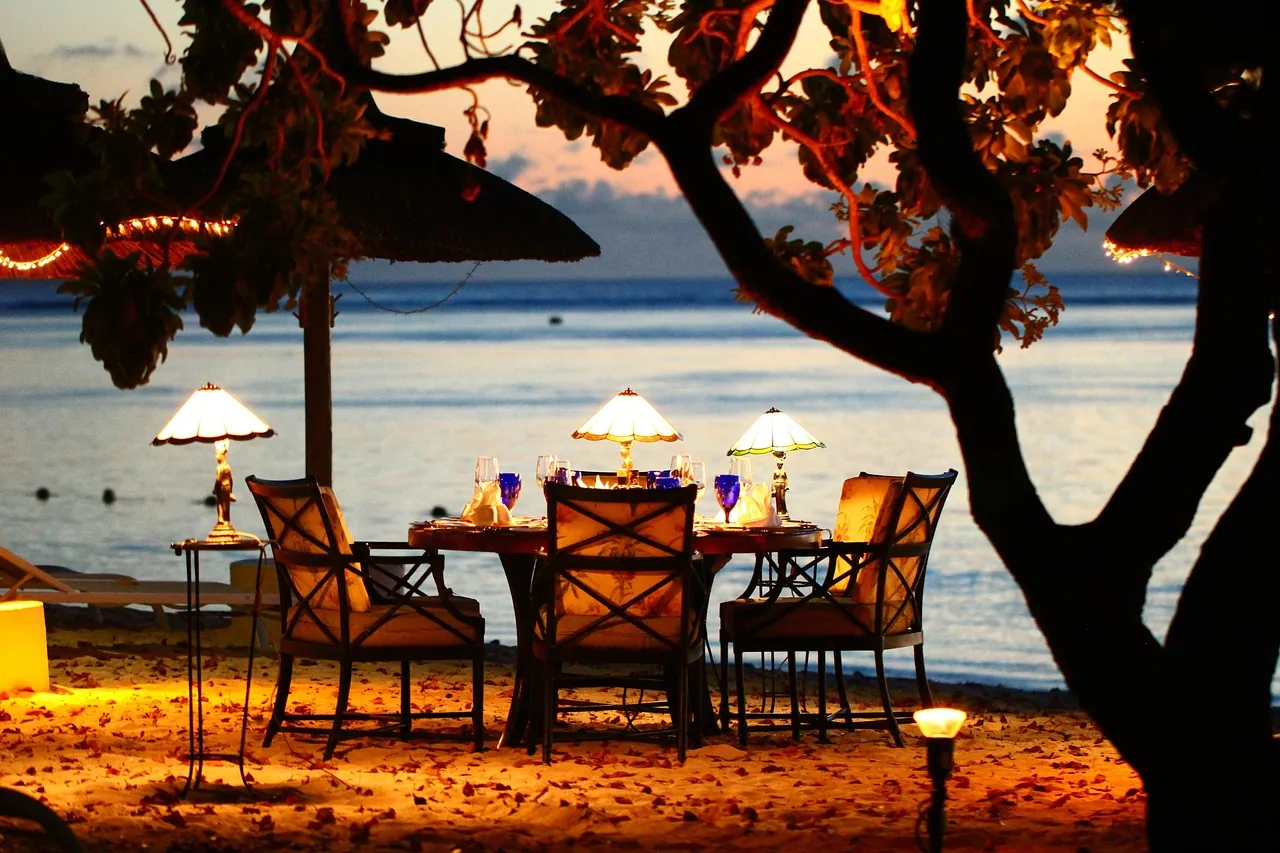 A dinner table set on the beach at dusk beneath a tree