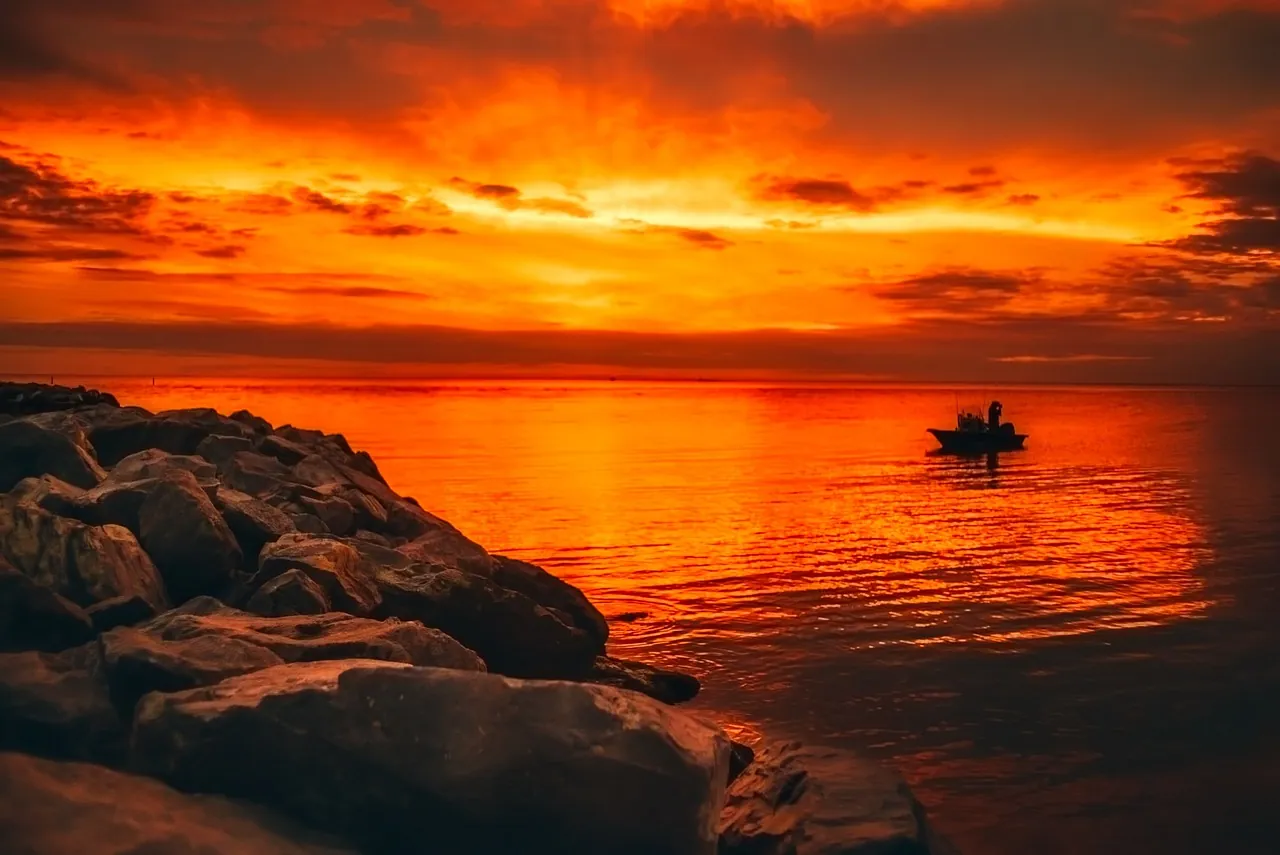 A small boat on glowing orange water beside dark rocks at sunset