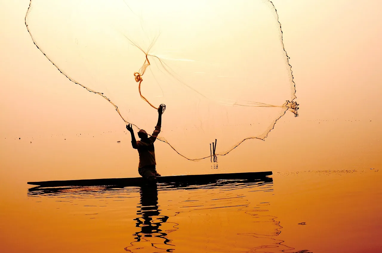 A fisherman casting a net from a small boat at sunrise